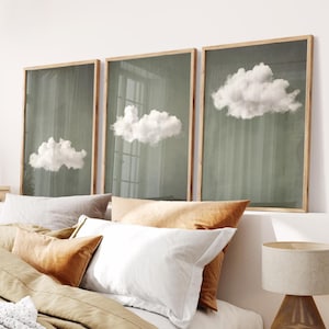 Puede incluir: Tres impresiones artísticas enmarcadas con nubes blancas y esponjosas sobre un fondo verde apagado. Los marcos tienen un acabado de madera natural. Las impresiones se exhiben sobre una cama con almohadas en tonos blancos, beige y naranja.
