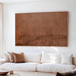 Può includere: Una grande opera d'arte murale rettangolare caratterizzata da una superficie testurizzata color marrone ruggine monocromatico con sottili motivi a goccia verticali, racchiusa in una sottile cornice di legno chiaro, esposta sopra un divano bianco.