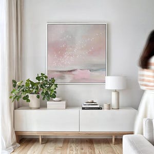 May include: A white media console with two drawers and a light wood base. A potted plant, a white box, and a stack of books are on top of the console. A white lamp with a white shade is on the right side of the console. A framed abstract painting with a pink and gray color scheme hangs on the wall above the console. The painting depicts a flock of white birds flying over a pink and gray landscape.