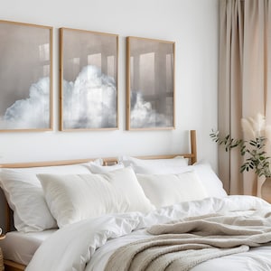 Puede incluir: Un dormitorio con una cama blanca, una manta beige y tres impresiones enmarcadas de nubes en una pared blanca. La cama tiene una cabecera de madera y almohadas blancas. Hay un jarrón con hierba de la pampa seca y una planta en maceta en una mesa auxiliar.