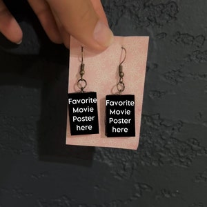 May include: A pair of black dangle earrings with gold hooks. The earrings feature a rectangular charm with the text "Favorite Movie Poster here" printed in white.