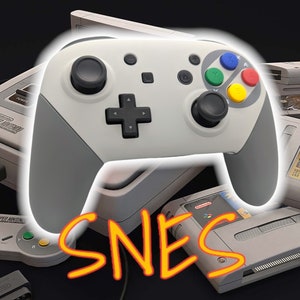 May include: A gray and white video game controller with colorful buttons, a Super Nintendo Entertainment System (SNES) console, and a game cartridge. The controller is a modern design with a d-pad and analog sticks.