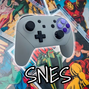 May include: A gray Nintendo Switch controller with purple buttons and a black faceplate. The controller is on top of a stack of comic books with the word "SNES" written in black and white.