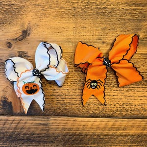 May include: Two Halloween-themed hair bows. One is white with a pumpkin design, the other is orange with a spider design. Both bows have black trim and a black and white polka dot centre.