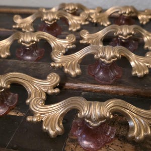 May include: A collection of six ornate drawer pulls featuring a metallic gold finish with intricate scrollwork and curved edges. Each handle is mounted on a translucent, deep reddish-brown base that flares outward at the bottom attachment point.
