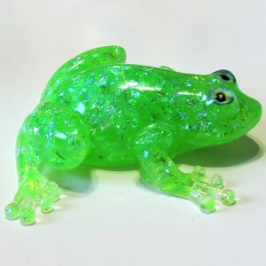 May include: A bright green frog toy with glitter embedded in the plastic. The frog has blue eyes and a textured surface.
