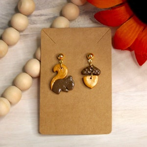 May include: A pair of earrings featuring a brown and gold squirrel and a brown and gold acorn. The earrings are displayed on a brown card.