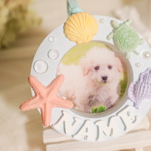 Pode incluir: Moldura de foto de cerâmica branca com um design de concha, estrela do mar e tartaruga. A moldura tem uma abertura redonda com uma foto de um cão branco dentro. A moldura diz "NAME" na parte inferior.