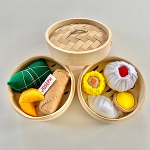 May include: A set of three bamboo steamer baskets filled with felt food items, including a green dumpling, a brown spring roll, a yellow fortune cookie, a yellow steamed bun with red accents, a white steamed bun with a red dot, a white dumpling, and a yellow custard tart. The basket on the left has a white tag with the name "Joseph" written on it.