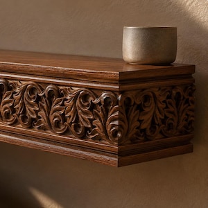 Pu&ograve; includere: Una mensola galleggiante in legno marrone scuro con un intricato motivo a foglie intagliato lungo il bordo anteriore. Una piccola ciotola in ceramica di colore chiaro &egrave; appoggiata sulla superficie superiore. La mensola mostra il logo MasterOak Since 1998.
