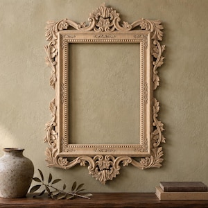 Puede incluir: Un marco de fotos rectangular de madera de color marrón claro con intrincados detalles tallados de motivos florales y volutas en los bordes, un borde interior de cuentas y un adorno decorativo ornamentado en el centro superior e inferior.