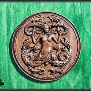 Op de afbeelding: Een houten wandplaquette met daarop een vrouw met slangenhaar, omringd door vier zeeslangen. De plaquette is rond en heeft een donkerbruine afwerking.