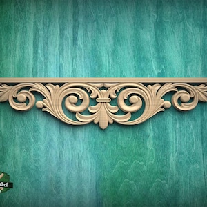 May include: A light-colored wooden decorative molding featuring an intricate carved scrollwork pattern with symmetrical floral-inspired motifs and a horizontal top edge. The piece displays a natural wood grain texture and detailed relief carvings.