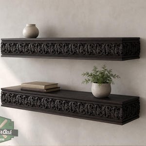 Pu&ograve; includere: Due mensole galleggianti in legno marrone scuro montate su una parete. Ogni mensola presenta un dettagliato motivo a foglie intagliate lungo il bordo anteriore. Un piccolo vaso in ceramica beige si trova sulla mensola superiore, mentre una pila di due libri e una piccola pianta in vaso si trovano sulla mensola inferiore. Il logo Master Oak &egrave; visibile nell'angolo in basso a sinistra.
