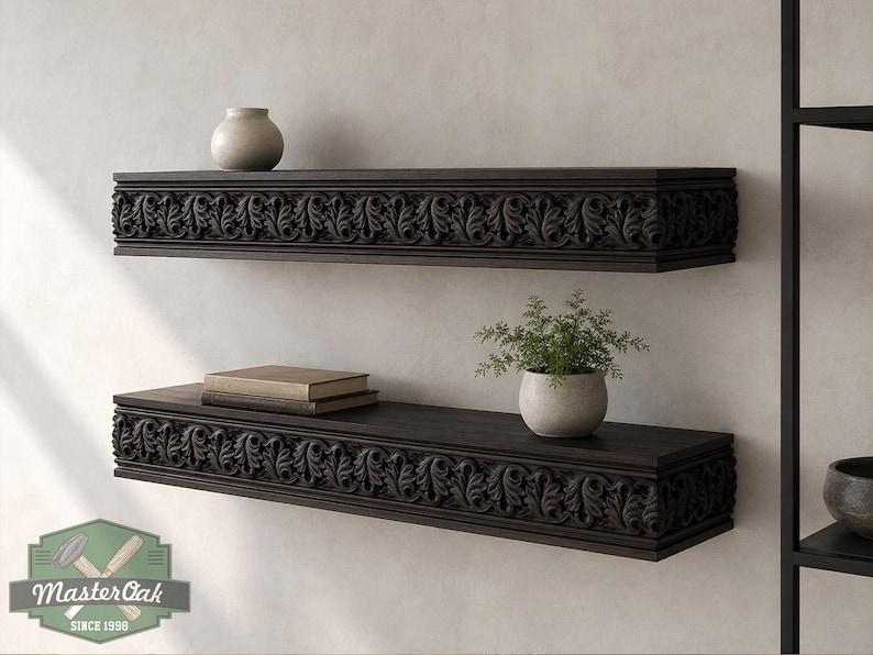 Pu&ograve; includere: Due mensole galleggianti in legno marrone scuro montate su una parete. Ogni mensola presenta un dettagliato motivo a foglie intagliate lungo il bordo anteriore. Un piccolo vaso in ceramica beige si trova sulla mensola superiore, mentre una pila di due libri e una piccola pianta in vaso si trovano sulla mensola inferiore. Il logo Master Oak &egrave; visibile nell'angolo in basso a sinistra.