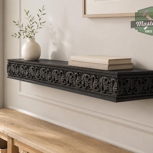 Pu&ograve; includere: Una mensola sospesa in legno marrone scuro con un intricato motivo a foglie intagliate lungo il bordo anteriore. La mensola sostiene un vaso in ceramica bianca con rametti verdi e due libri beige impilati. Un logo nell'angolo riporta MasterOak Since 1998.