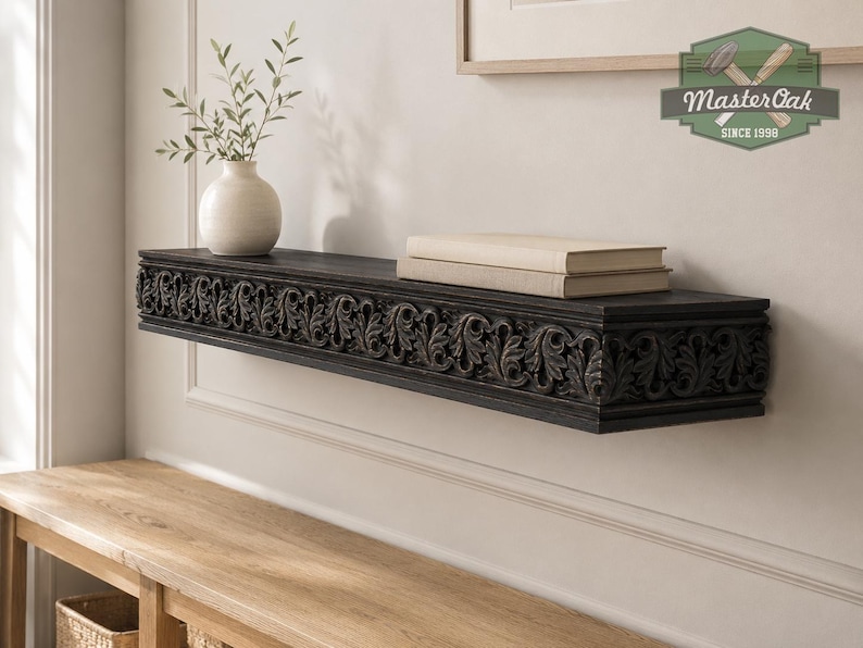 Pu&ograve; includere: Una mensola sospesa in legno marrone scuro con un intricato motivo a foglie intagliate lungo il bordo anteriore. La mensola sostiene un vaso in ceramica bianca con rametti verdi e due libri beige impilati. Un logo nell'angolo riporta MasterOak Since 1998.