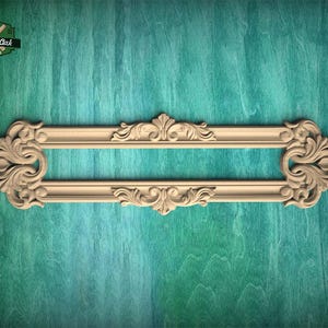 Puede incluir: Una moldura decorativa de madera clara con un marco rectangular alargado que presenta intrincados tallados de volutas y motivos florales en los extremos y secciones centrales. La pieza muestra una textura de veta de madera natural y detalles en relieve simétricos.