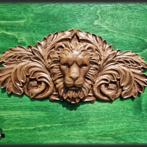 Puede incluir: Una cabeza de león tallada en madera con detalles de hojas ornamentadas, montada sobre un fondo verde.
