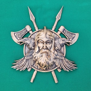 Puede incluir: Una decoración de pared de madera con un guerrero vikingo tallado con barba, que lleva un casco y está rodeado de dos cuervos y hachas cruzadas. La cara del guerrero es severa y sus ojos son penetrantes. El fondo es una tela verde.