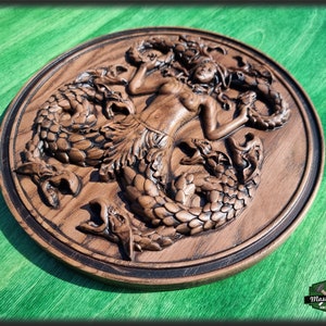 Op de afbeelding: Een houten wandplaquette met een gedetailleerde carving van een zeemeermin omringd door drie zeeslangen. De plaquette is rond en heeft een donkerbruine vlek.