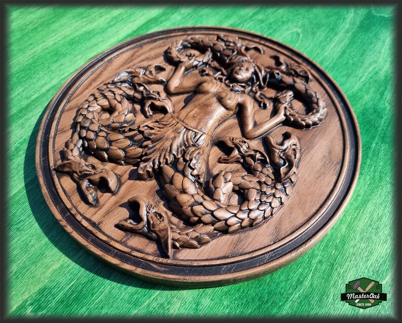 Op de afbeelding: Een houten wandplaquette met een gedetailleerde carving van een zeemeermin omringd door drie zeeslangen. De plaquette is rond en heeft een donkerbruine vlek.