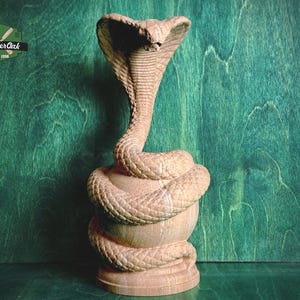 Op de afbeelding: Houten cobra sculptuur met gedetailleerde gravures. De slang is opgerold rond een basis, met zijn kop omhoog. De sculptuur is gemaakt van lichtgekleurd hout en staat tegen een groene achtergrond. Het logo "Master Oak Since 1996" is zichtbaar.
