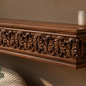 Pu&ograve; includere: Una mensola galleggiante in legno marrone scuro con un intricato motivo a foglie intagliato lungo il bordo anteriore. Una piccola ciotola in ceramica di colore chiaro &egrave; appoggiata sulla superficie superiore. La mensola mostra il logo MasterOak Since 1998.