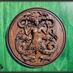 Op de afbeelding: Een bruine houten wandplaquette met een gegraveerd beeld van een vrouw met slangen als haar en een staart. De vrouw is omringd door slangen en heeft een woeste uitdrukking op haar gezicht.