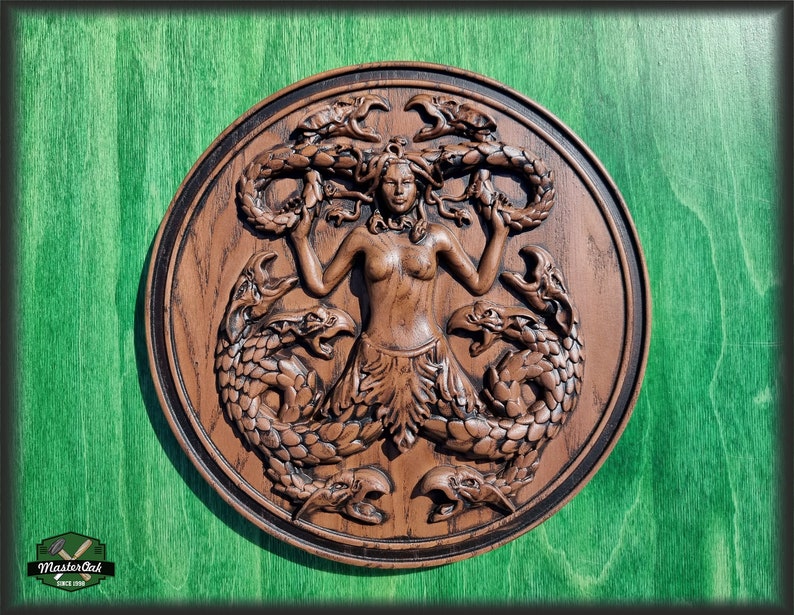 Op de afbeelding: Een bruine houten wandplaquette met een gegraveerd beeld van een vrouw met slangen als haar en een staart. De vrouw is omringd door slangen en heeft een woeste uitdrukking op haar gezicht.