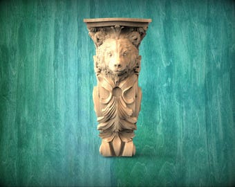 May include: A carved wooden architectural element featuring a bear's head. The light brown piece has detailed carvings of the bear's face, fur, and decorative foliage. The piece is approximately 30 cm tall.