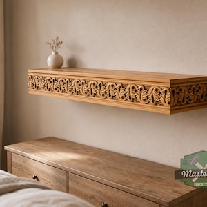 Pu&ograve; includere: Una mensola galleggiante in legno marrone chiaro con un intricato motivo a foglie intagliate sul bordo anteriore. Un piccolo vaso bianco con fiori secchi si trova sul lato sinistro. Il testo MasterOak SINCE 1998 &egrave; visibile nell'angolo in basso a destra.