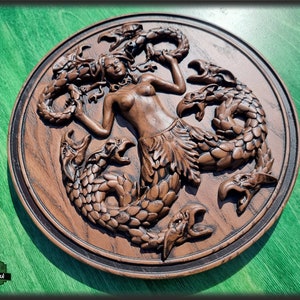Op de afbeelding: Een houten wandplaquette met een gedetailleerde carving van een zeemeermin omringd door drie draken. De zeemeermin is afgebeeld met lang, golvend haar en een staart, terwijl de draken schubben en vleugels hebben. De plaquette is geschilderd in tinten bruin en zwart.