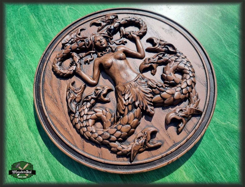 Op de afbeelding: Een houten wandplaquette met een gedetailleerde carving van een zeemeermin omringd door drie draken. De zeemeermin is afgebeeld met lang, golvend haar en een staart, terwijl de draken schubben en vleugels hebben. De plaquette is geschilderd in tinten bruin en zwart.