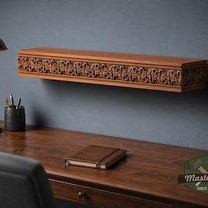 Pu&ograve; includere: Una mensola rettangolare in legno con un intricato motivo a foglie intagliato sul bordo anteriore. La mensola &egrave; montata a parete sopra una scrivania in legno, sulla quale si trovano una lampada in ottone, un portapenne e un taccuino chiuso.