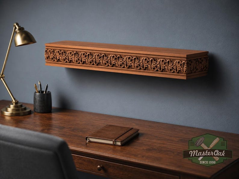 Pu&ograve; includere: Una mensola rettangolare in legno con un intricato motivo a foglie intagliato sul bordo anteriore. La mensola &egrave; montata a parete sopra una scrivania in legno, sulla quale si trovano una lampada in ottone, un portapenne e un taccuino chiuso.
