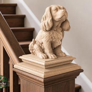 May include: A light-colored wooden sculpture of a spaniel dog sitting upright, featuring detailed fur texture and a raised head. The carving is mounted on a square tiered base atop a dark wood staircase newel post with a MasterOak logo visible.