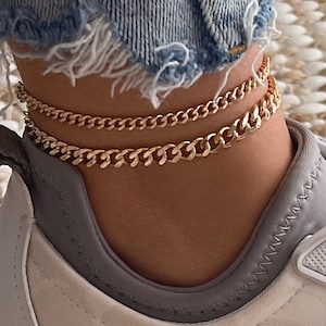 May include: A pair of gold ankle chains on a person's ankle wearing white sneakers with a gray trim.