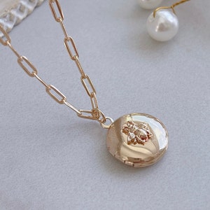 May include: Gold locket necklace with a delicate chain. The round locket features a detailed bee design. The necklace is displayed on a light grey surface, with decorative elements in the background.