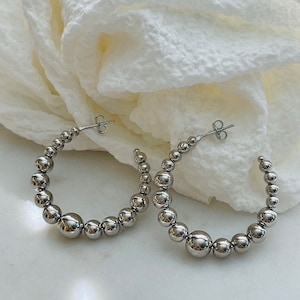 May include: A pair of silver hoop earrings with a beaded design. The earrings are made of small, round beads that are connected together to form a hoop shape.