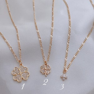 May include: Three gold necklaces with different charms. The first necklace has a four-leaf clover charm with clear rhinestones. The second necklace has a smaller four-leaf clover charm with clear rhinestones. The third necklace has a small cluster of clear rhinestones.