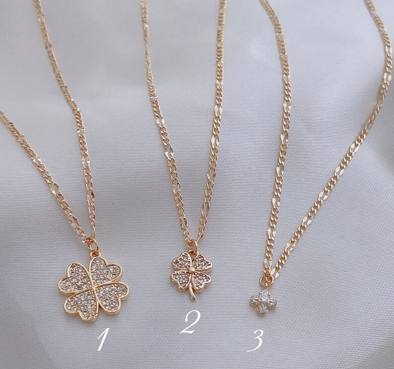 May include: Three gold necklaces with different charms. The first necklace has a four-leaf clover charm with clear rhinestones. The second necklace has a smaller four-leaf clover charm with clear rhinestones. The third necklace has a small cluster of clear rhinestones.