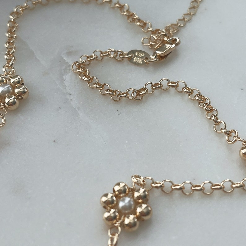 May include: A gold chain necklace with small flower charms. Each flower charm has a pearl in the centre. The chain has a clasp with the inscription "GF 18K".