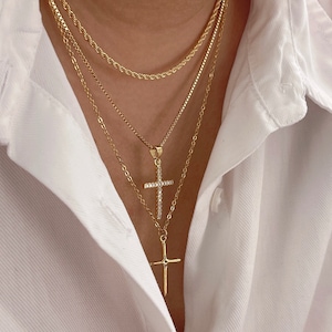 May include: Layered gold chain necklace with two cross pendants, one with small clear stones. The necklace is worn on a person with a white shirt.