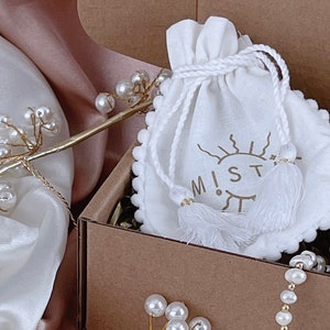 May include: A white drawstring pouch with a sun and eye design and the word "M!ST" printed on it. The pouch is in a brown cardboard box with white pearl necklaces.