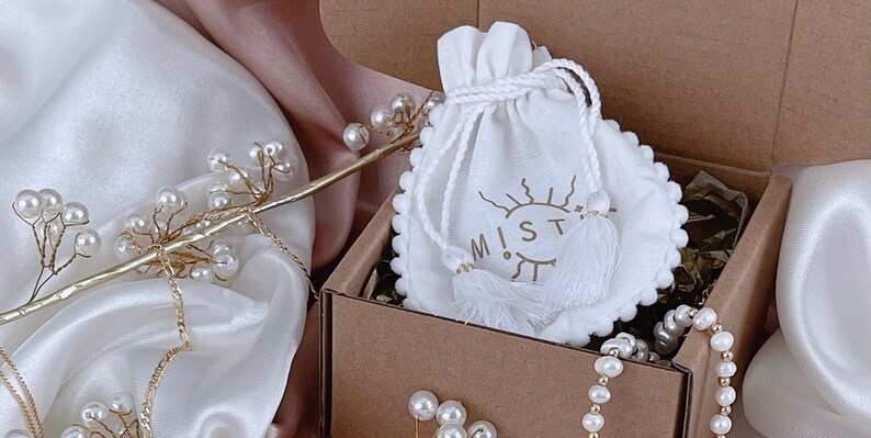 May include: A white drawstring pouch with a sun and eye design and the word "M!ST" printed on it. The pouch is in a brown cardboard box with white pearl necklaces.