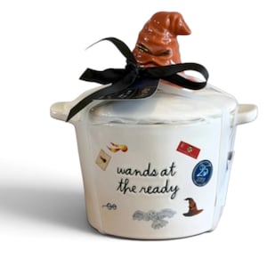 May include: A white ceramic lidded jar featuring a brown sculpted hat handle. The jar displays the text "wands at the ready" surrounded by small illustrations of an envelope, a red banner, a blue circular emblem, glasses, an owl, and a hat.