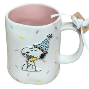 May include: White ceramic mug featuring Snoopy wearing a party hat and hugging Woodstock. The mug has a pink interior and is decorated with colorful confetti. A white ribbon and tag are attached to the handle.