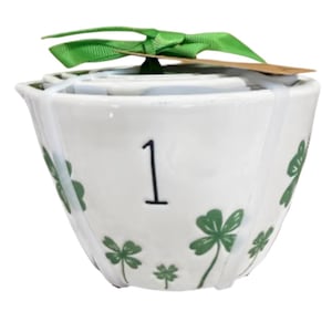 May include: White ceramic measuring cup set with green shamrock designs and the number 1 in black. The set is tied with a green ribbon and a small tag. A festive kitchenware item.