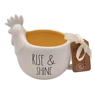 May include: White ceramic mug shaped like a rooster with a yellow interior. The mug has the words "RISE & SHINE" in black lettering. A brown tag is attached with a cream-colored ribbon.